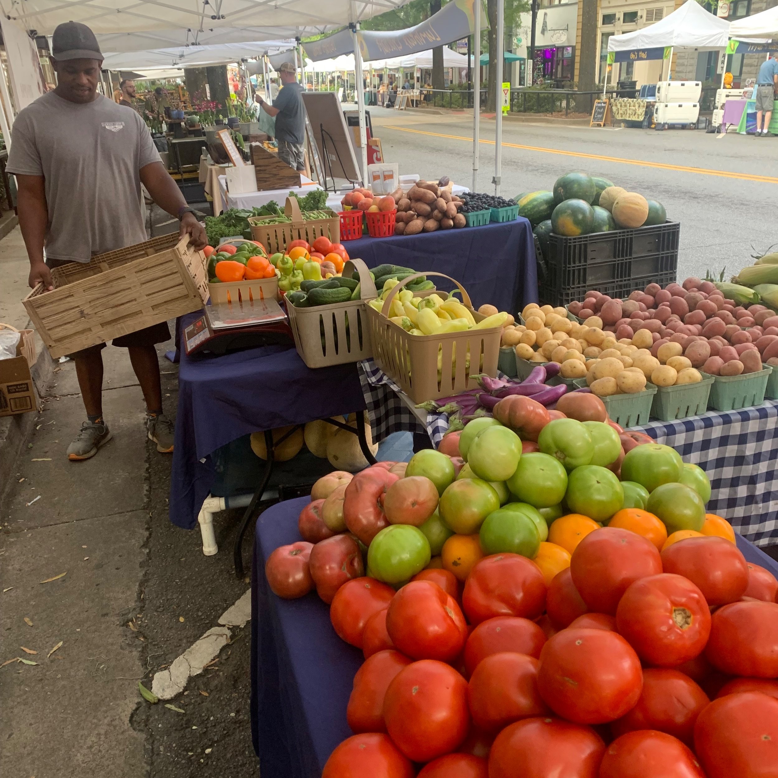 A vendor at a farmers market arranges fresh produce, including vibrant red tomatoes, green apples, and various vegetables, under white tents along a street.
