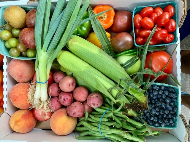 Fresh assortment of fruits and vegetables including peaches, potatoes, green beans, corn, cherry tomatoes, blueberries, and various peppers in a cardboard box, showcasing vibrant colors and healthy produce.